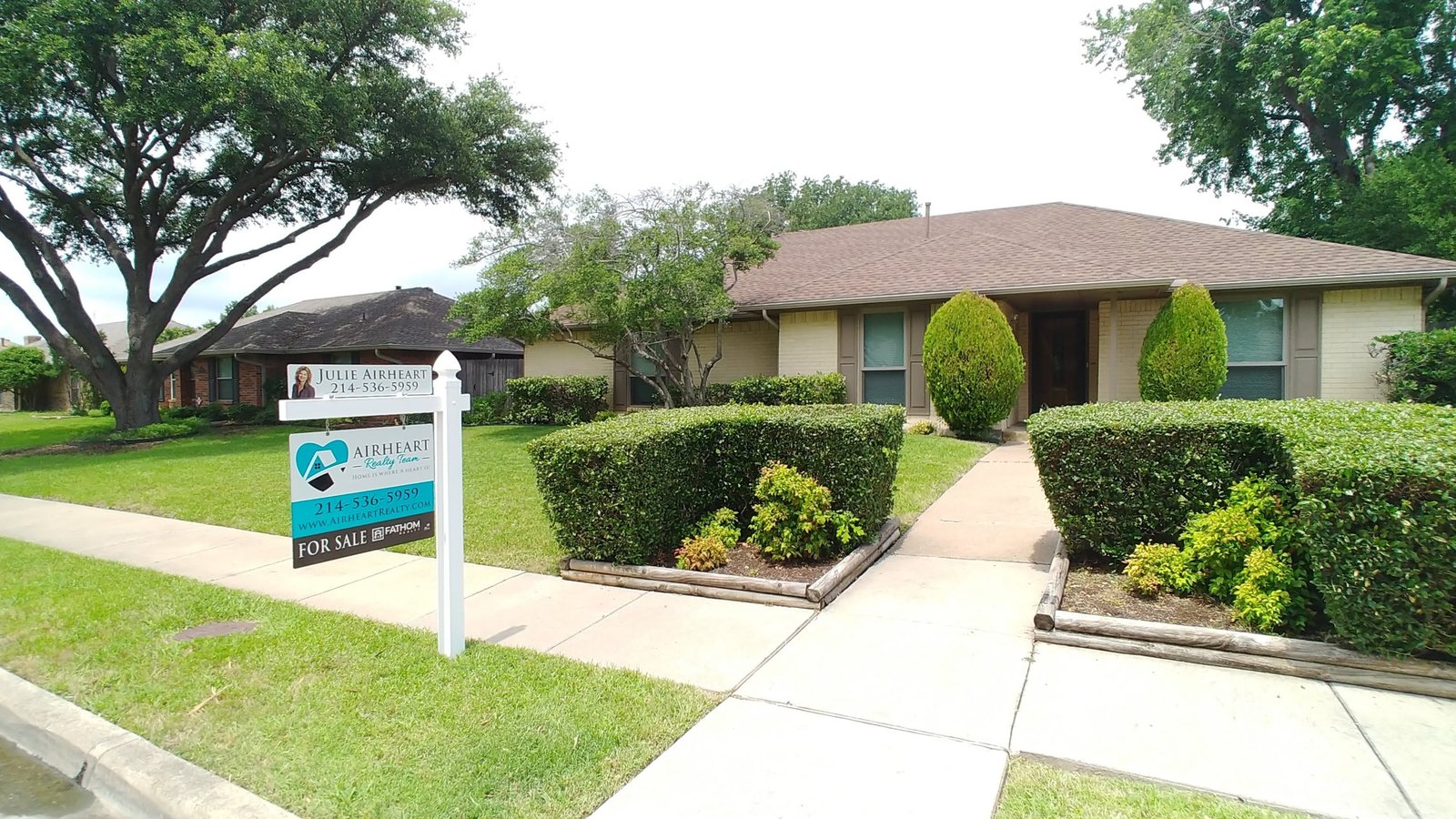 Airheart Realty Team For Sale sign in the front yard of a single-story brick home with trimmed hedges and mature oak trees