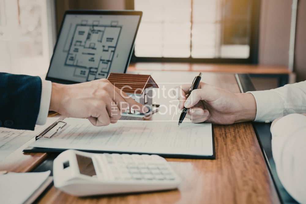 Two people review real estate paperwork beside a laptop showing a floor plan.