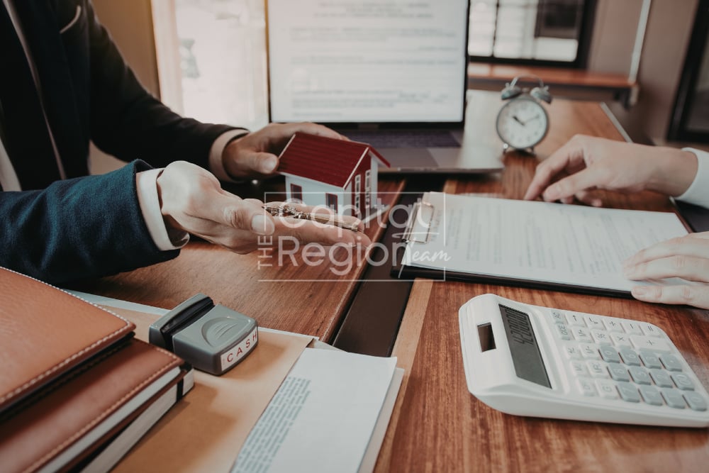 Hands exchange a model house over real estate paperwork on a desk.