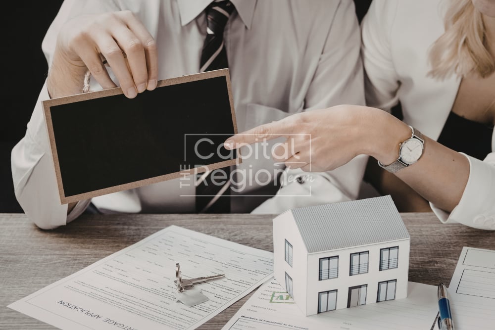 Two people review real estate paperwork beside a model house and blank sign.