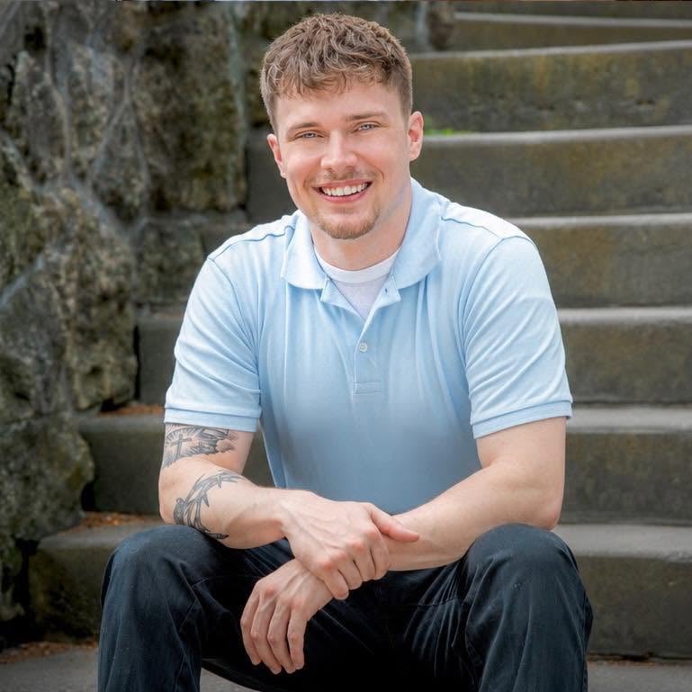 Kirk smiles in a light blue polo while seated on stone steps.