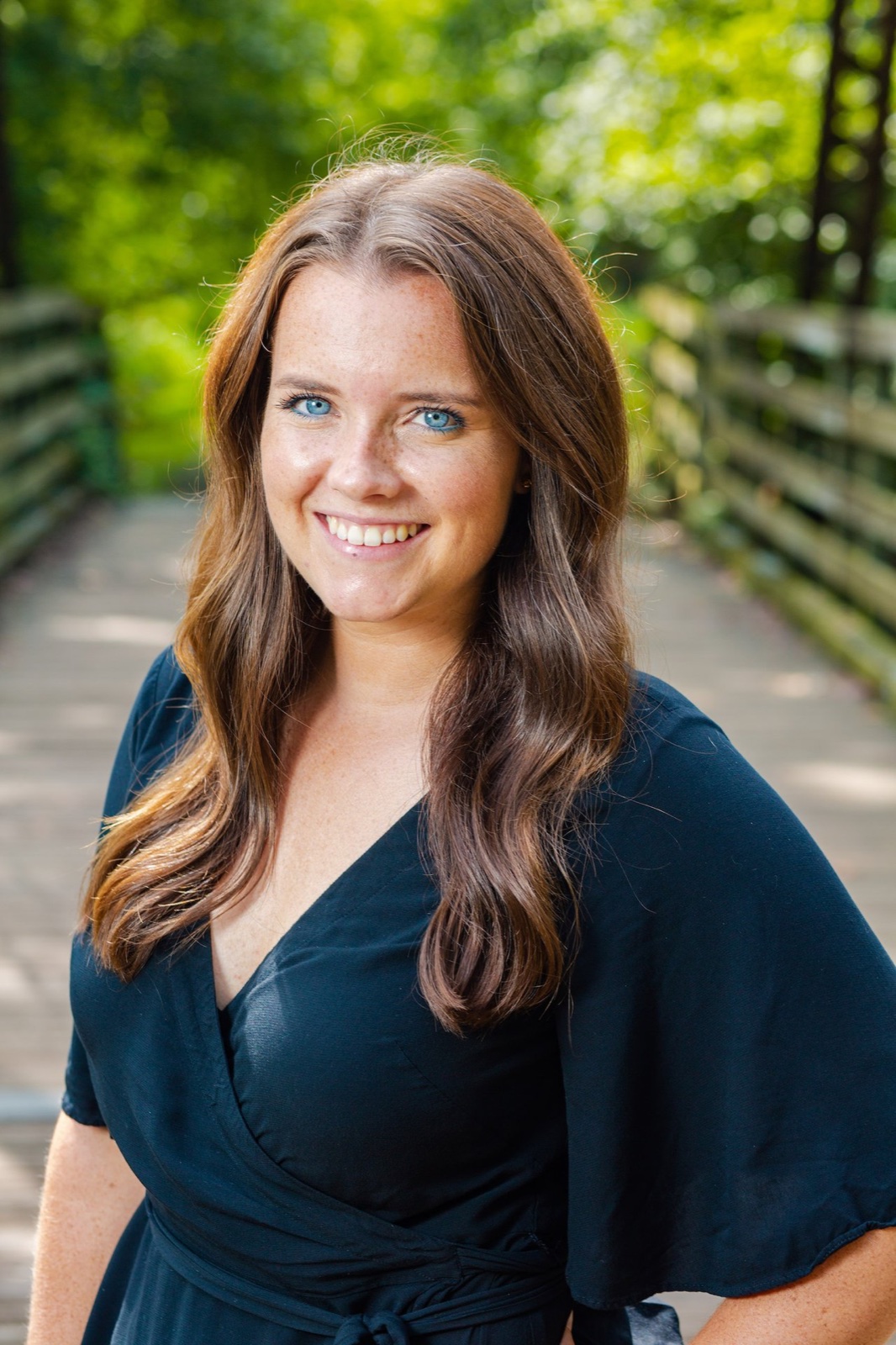 Reed, a smiling real estate agent with long brown hair, outdoors on a shaded walkway.