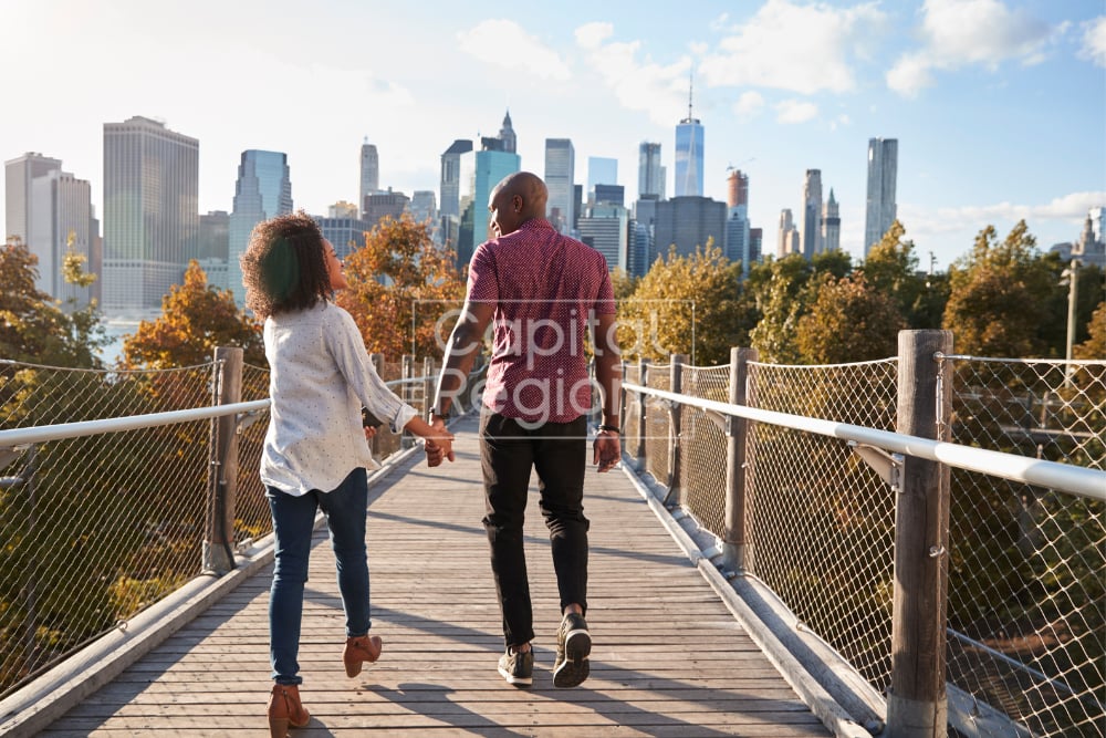Two people walk hand in hand across a bridge toward a city skyline.
