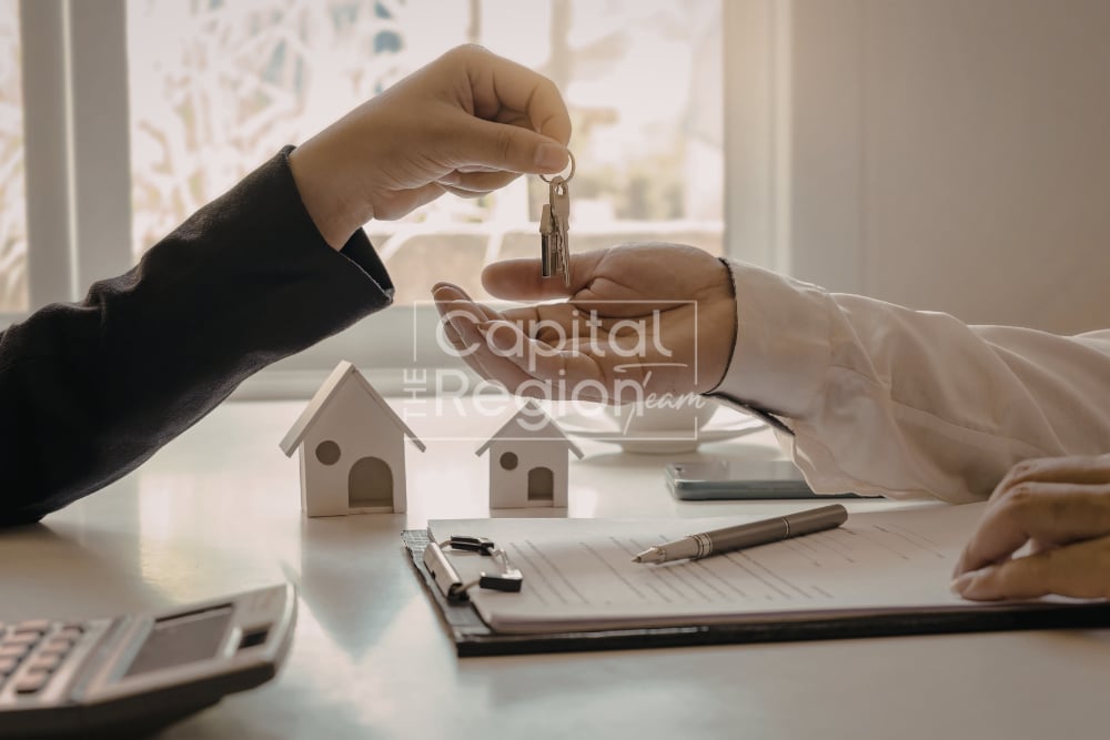 Two people exchange house keys over a desk with miniature house models and real estate paperwork.