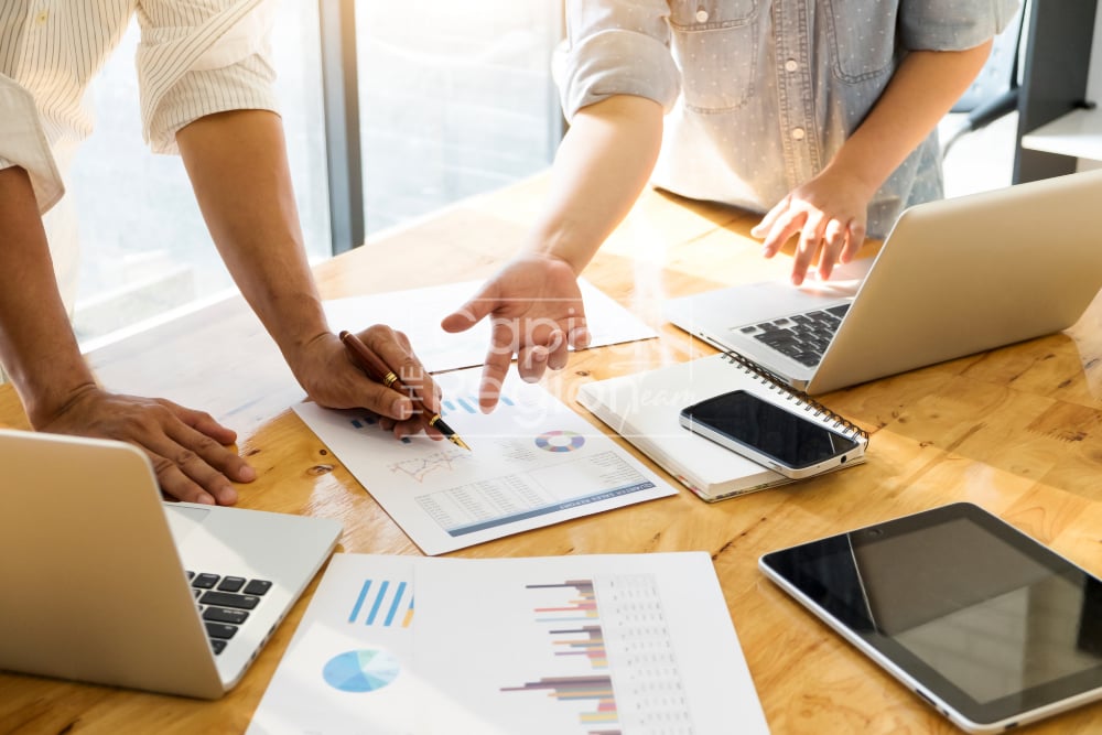 People review charts and documents together at a table with laptops and a smartphone.