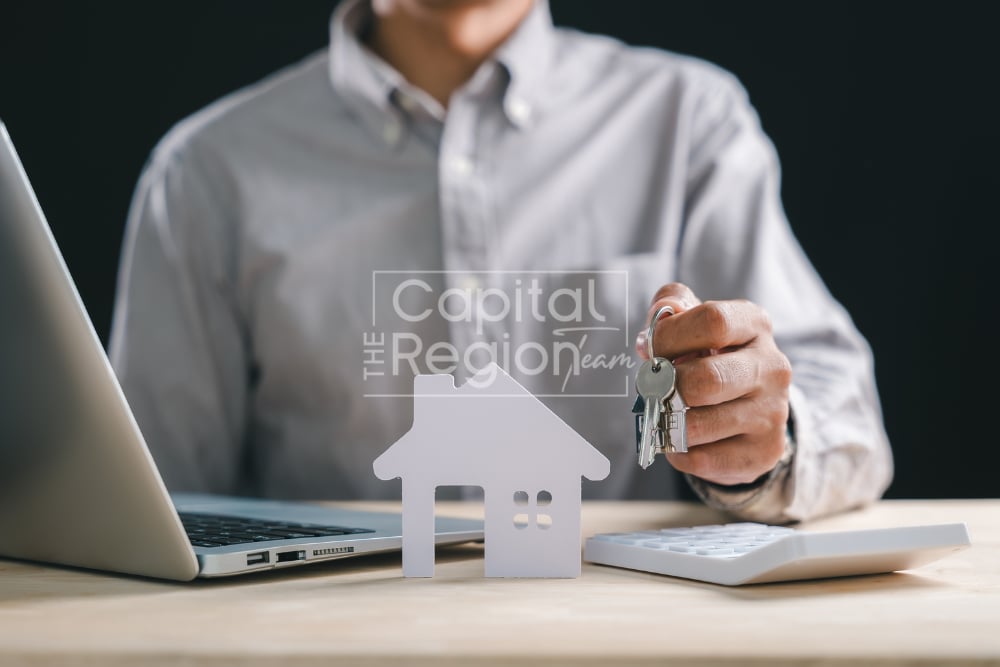 Person holding house keys beside a laptop, calculator, and house-shaped cutout.