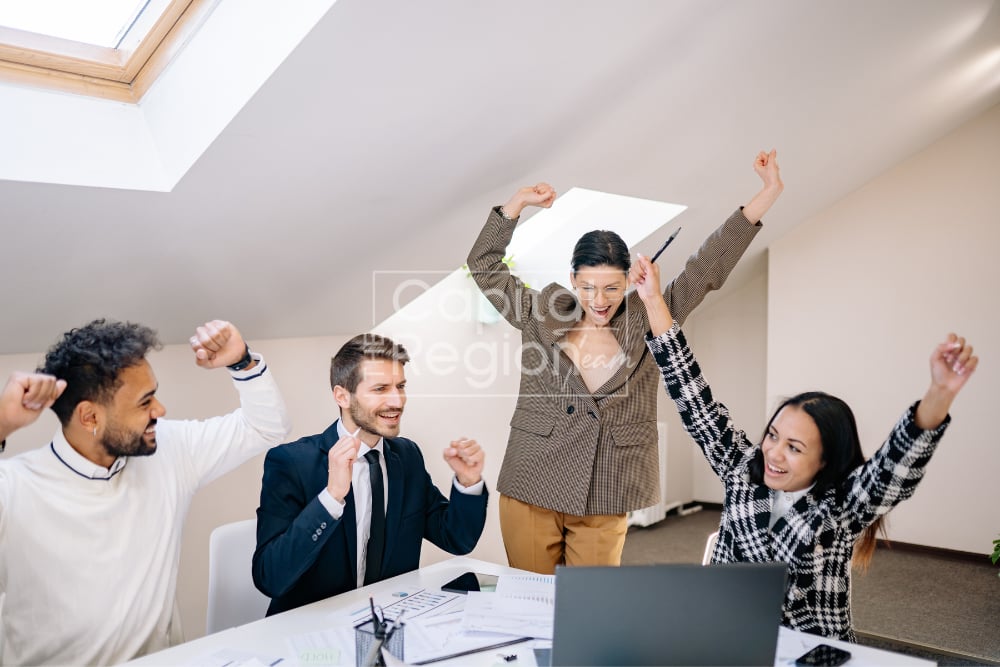 Four real estate professionals celebrate around a table with a laptop and paperwork.