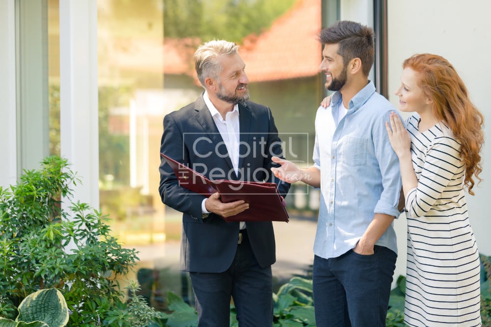 Real estate agent talks with a couple in a bright, glass-walled home entryway.
