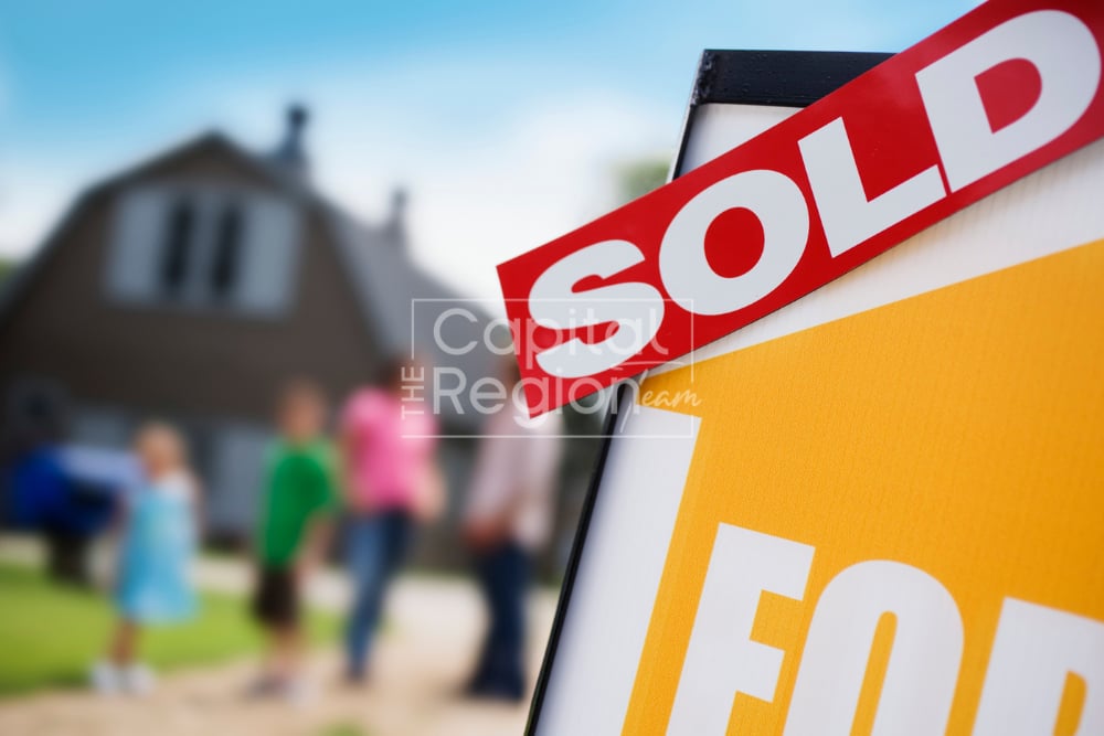 Real estate SOLD sign in front of a house, with a family viewing the property in the background.