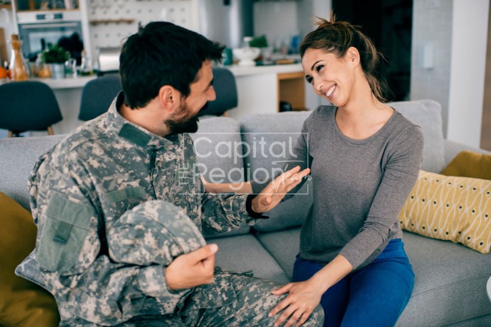 A smiling couple sits together on a sofa, the man wearing military fatigues.