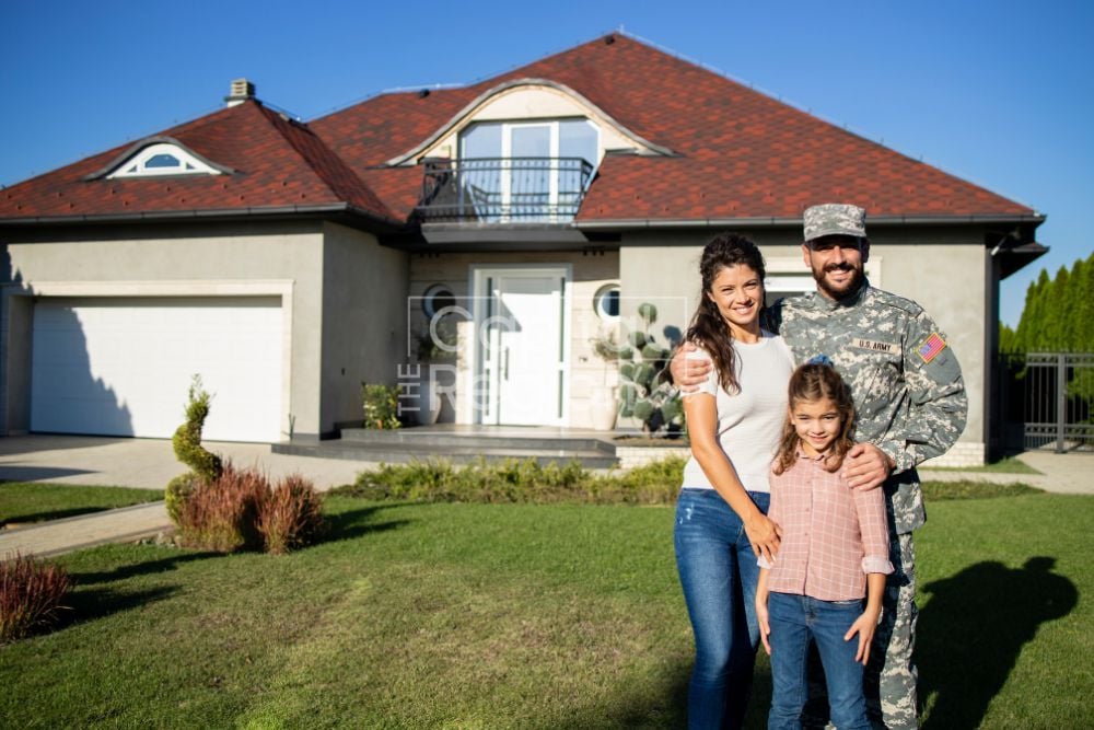 Family with a uniformed servicemember on the lawn in front of a two-story house with a red roof.