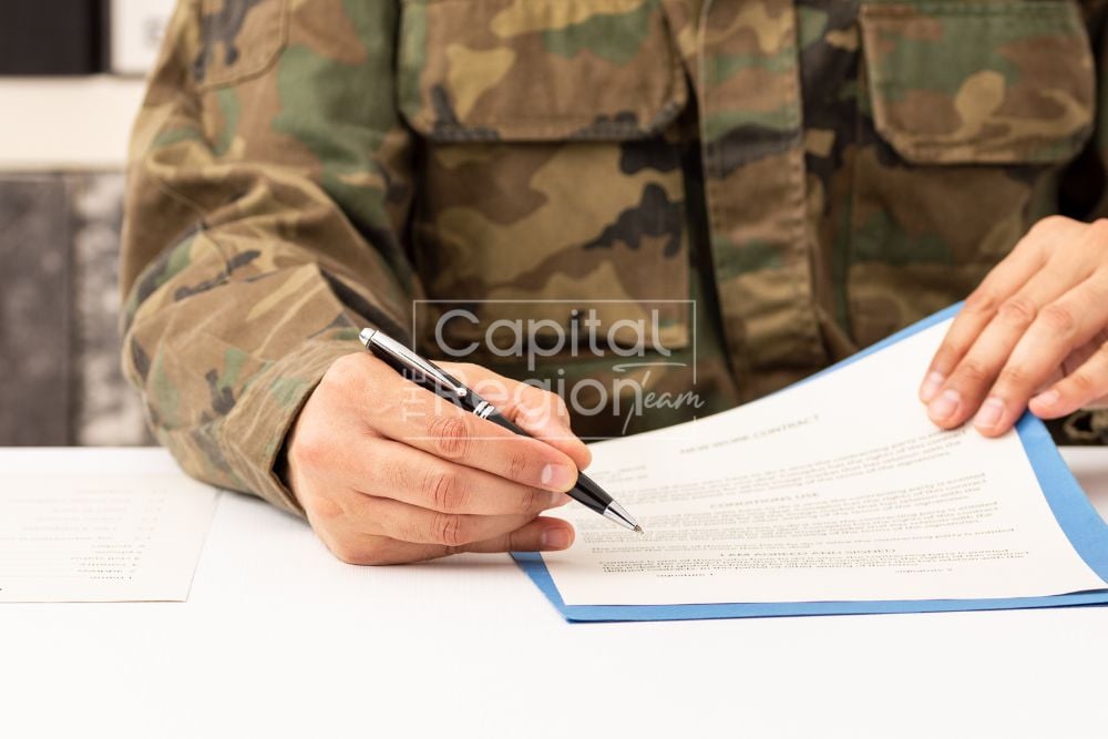 Service member in camouflage signs a document at a table.