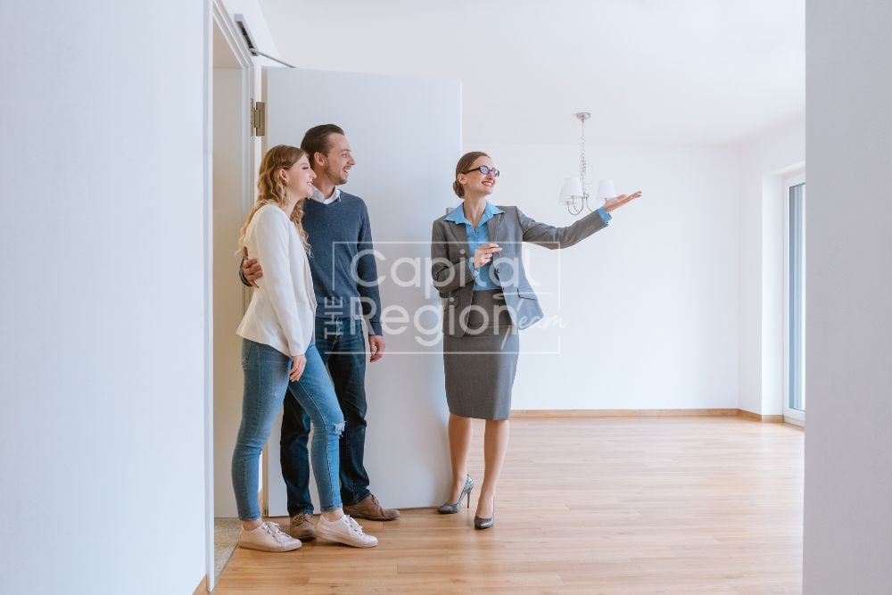 An agent shows a couple a bright, empty room with pale wood floors and a large window.