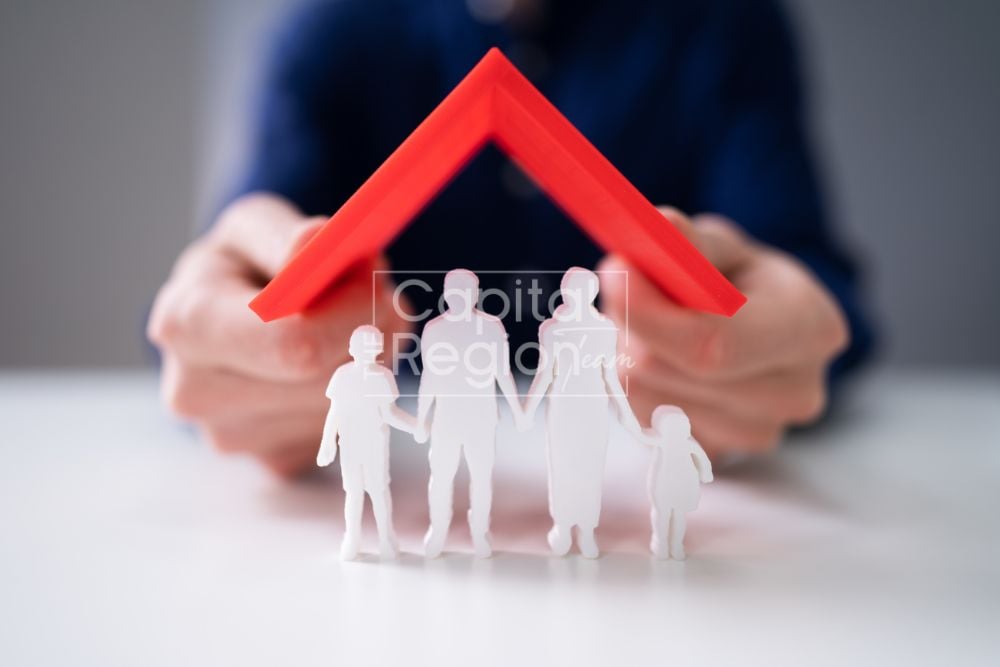Hands hold a red roof over white family figurines.