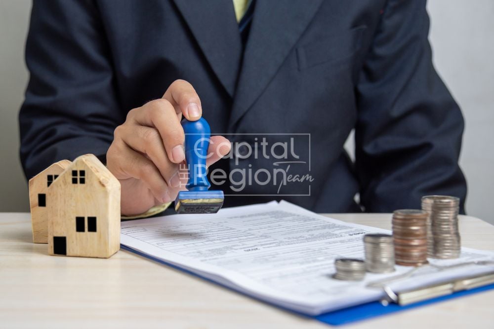 A hand stamps real estate paperwork beside a wooden house model and stacks of coins.