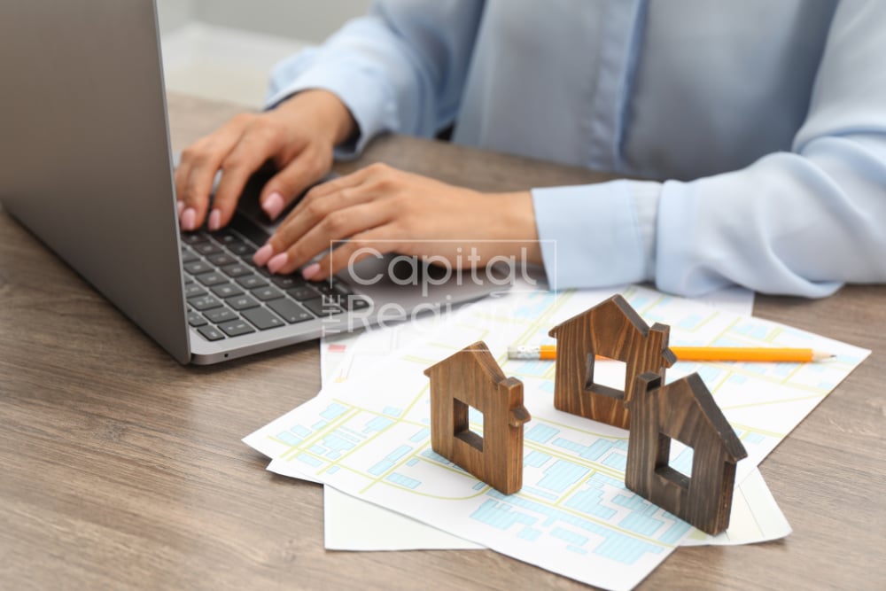 A person types on a laptop beside wooden house models and printed maps.