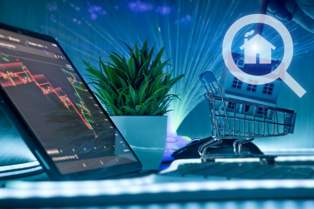 Tablet with a falling chart beside a miniature house in a shopping cart and magnifying-glass house symbol