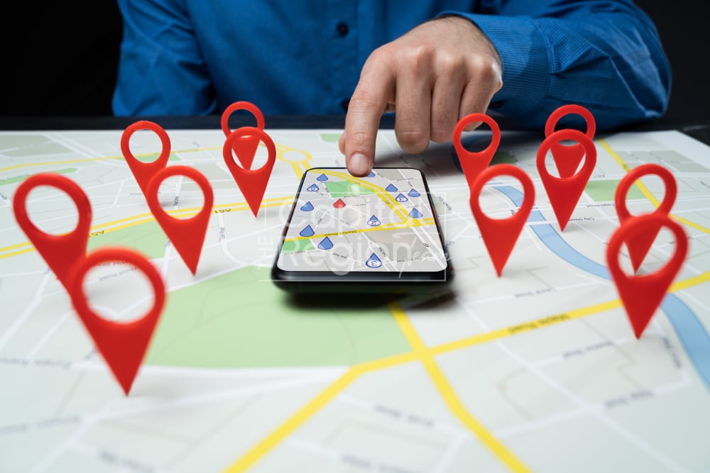 A hand selects a location on a smartphone map surrounded by red map pins.