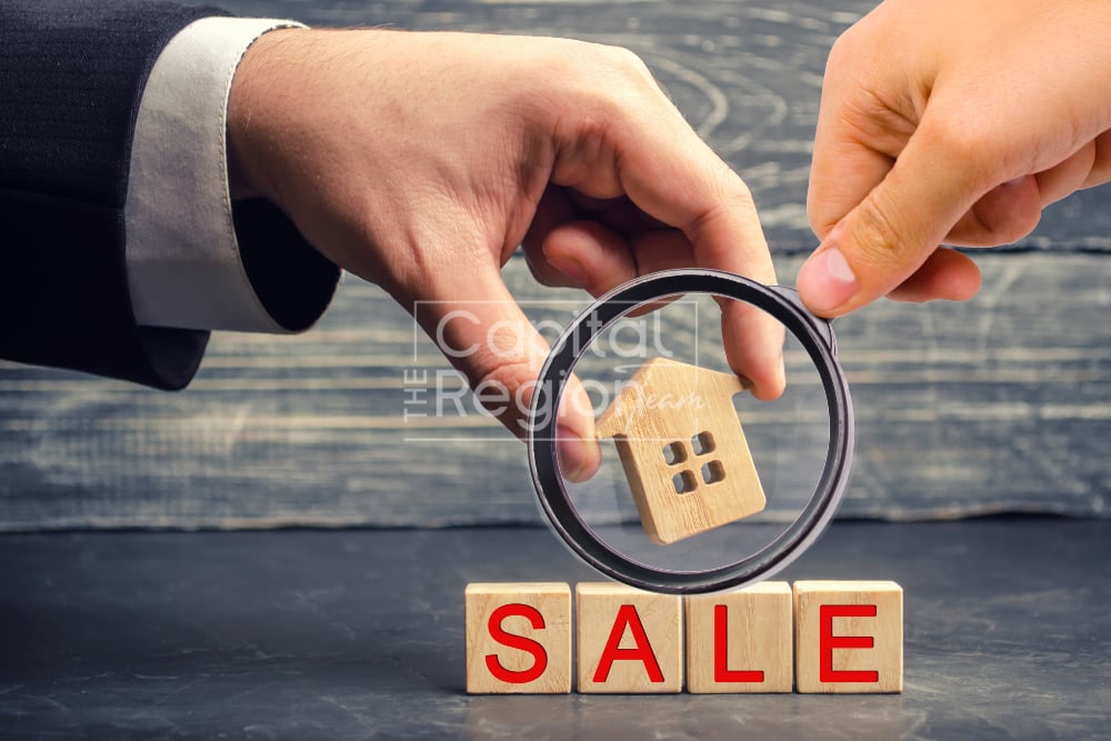 Hands inspect a wooden house with a magnifying glass above blocks spelling SALE.