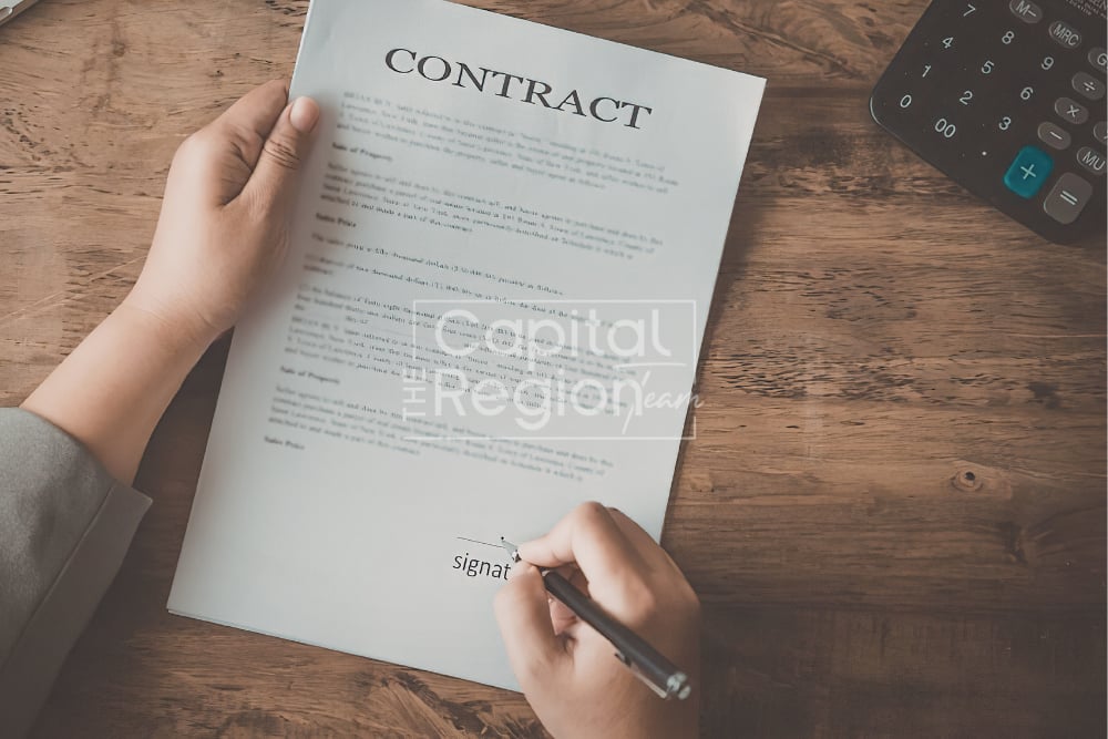Hands signing a home purchase contract beside a calculator on a wooden table.