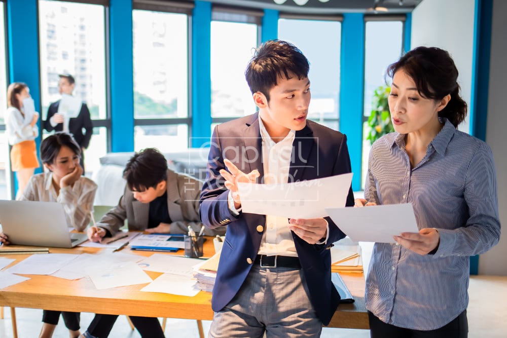 People review paperwork around a table in a bright office; two compare documents in the foreground.