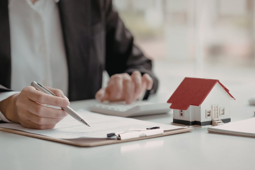 Person signing paperwork beside a model house while using a calculator.