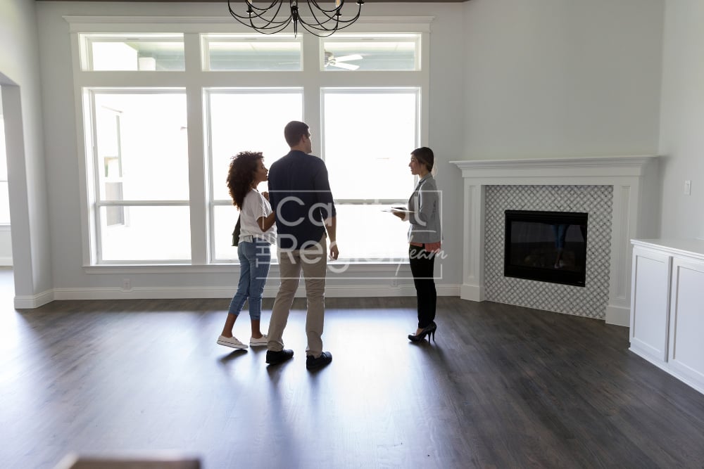 Unfurnished living room with large windows and a tiled fireplace, viewed by three people.