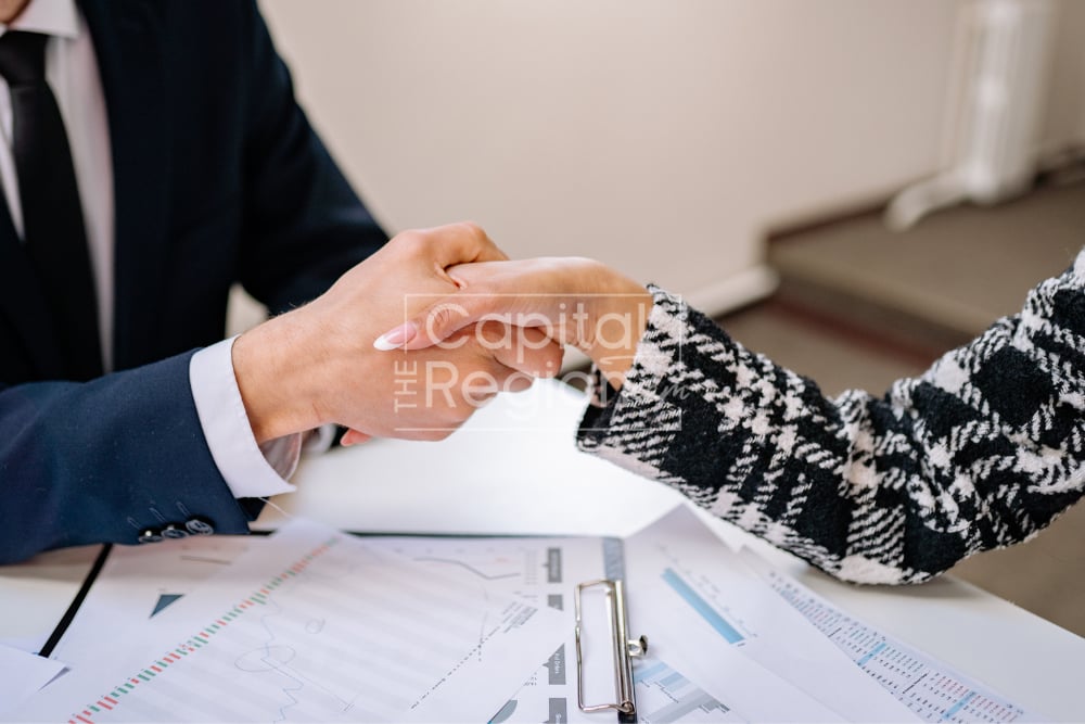 Two people shake hands over printed documents and charts at a table.