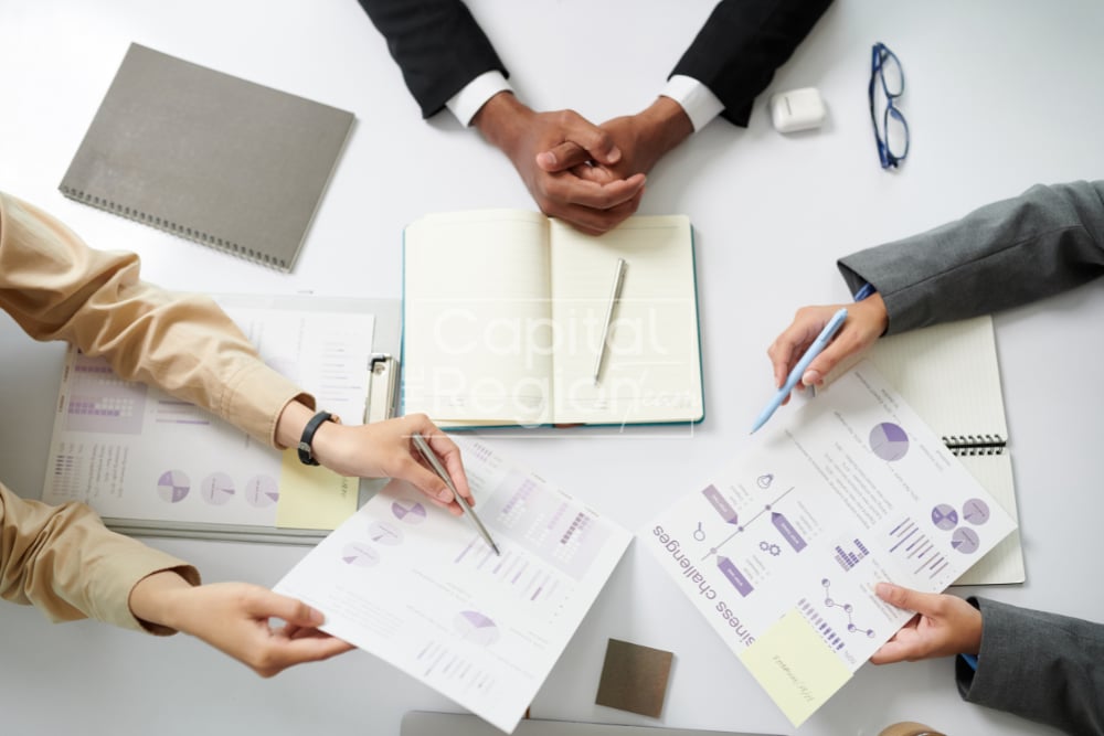 People review charts and reports around a table, with an open notebook and pens between them.