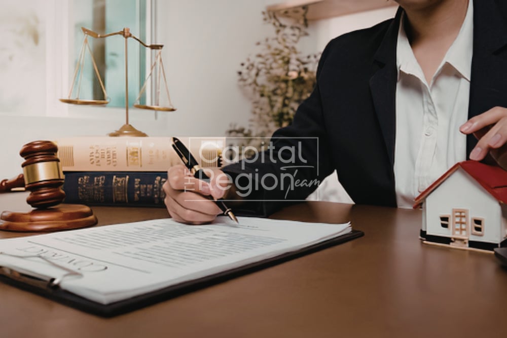 A person signs paperwork beside a model house, gavel, law books, and balance scales.
