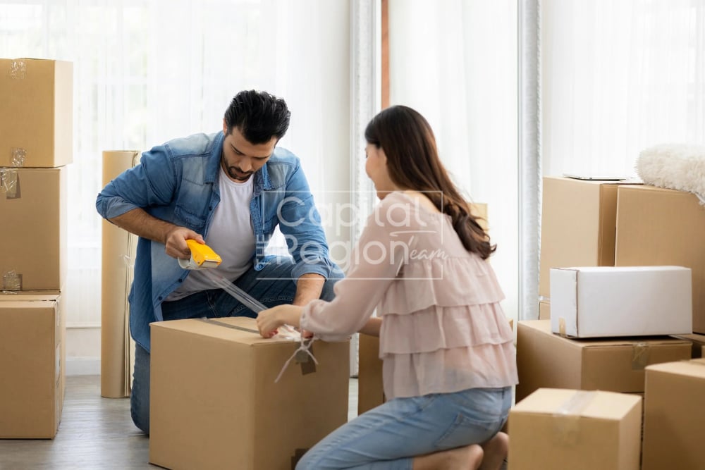 A couple packs a moving box together in a bright room surrounded by boxes.