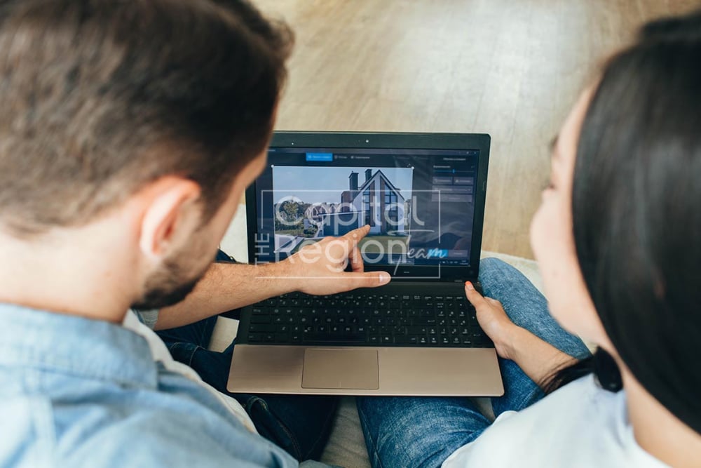 A couple browses home listings on a laptop, pointing at a house photo.
