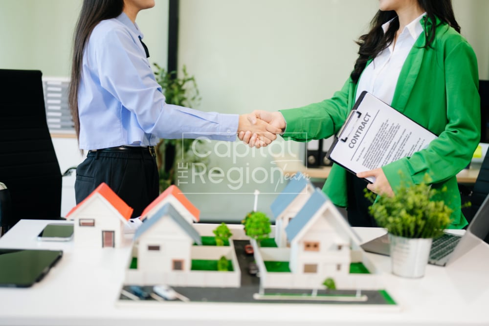 Real estate professionals shake hands over miniature house models, with one holding a contract.