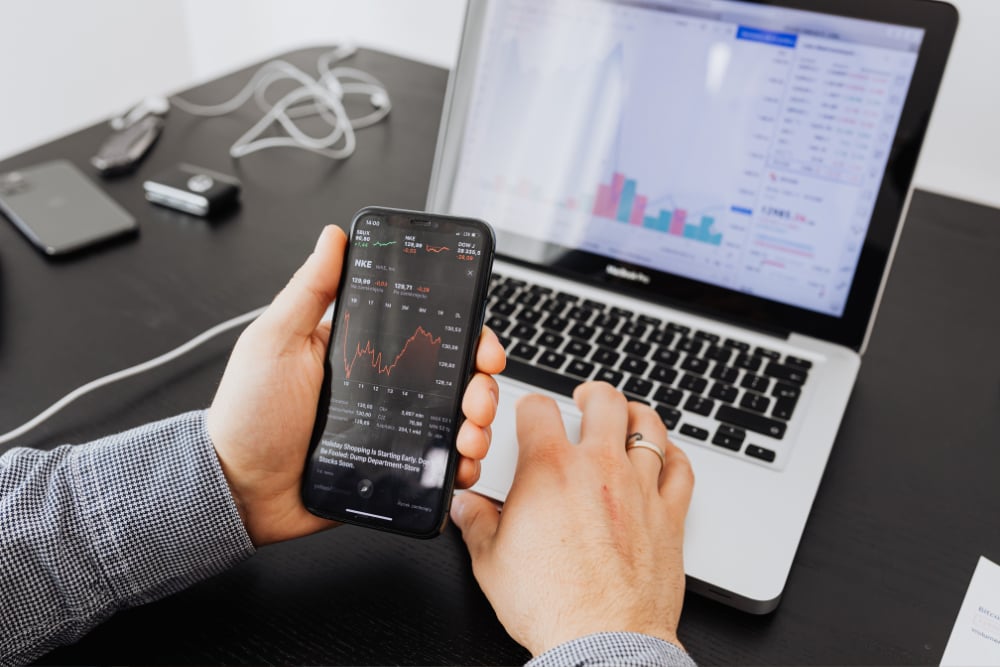 Person checks stock charts on a smartphone while using a laptop.