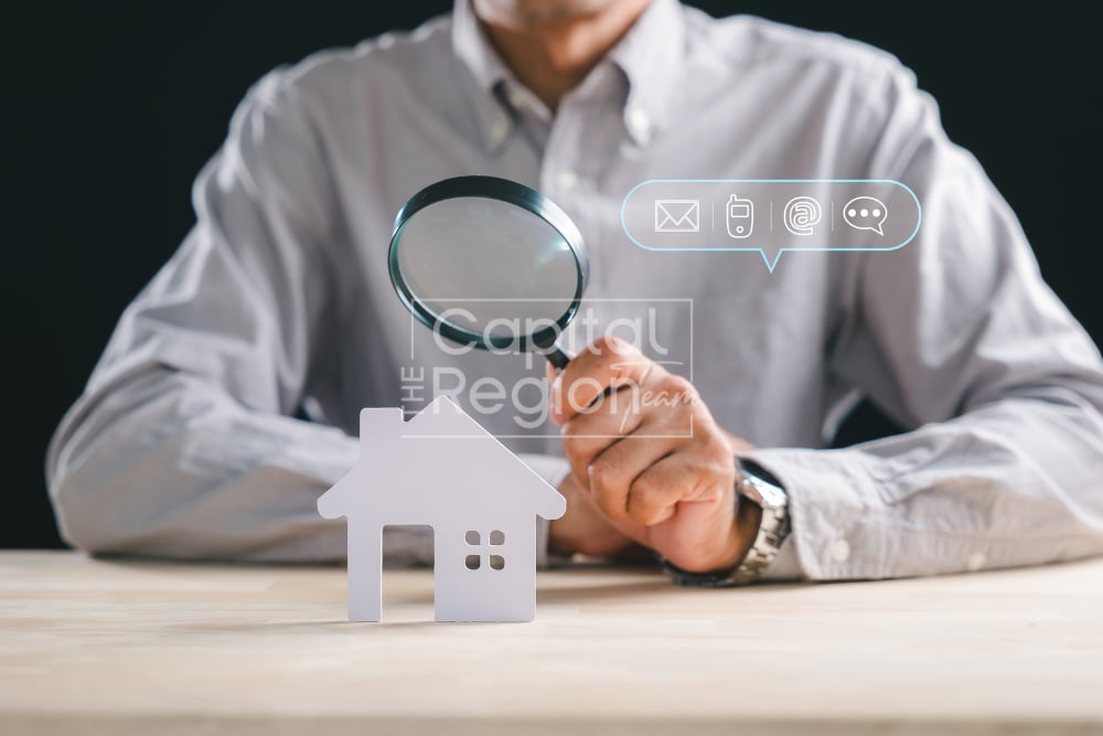 Person examining a paper house with a magnifying glass beside digital contact icons.