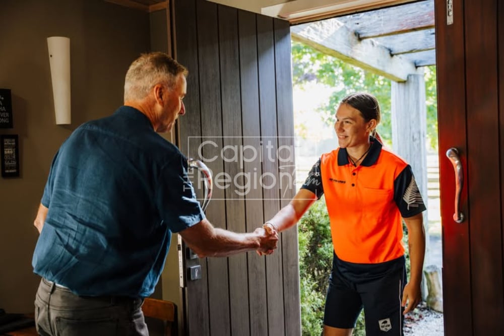 A man greets an orange-shirted visitor with a handshake at an open front door.