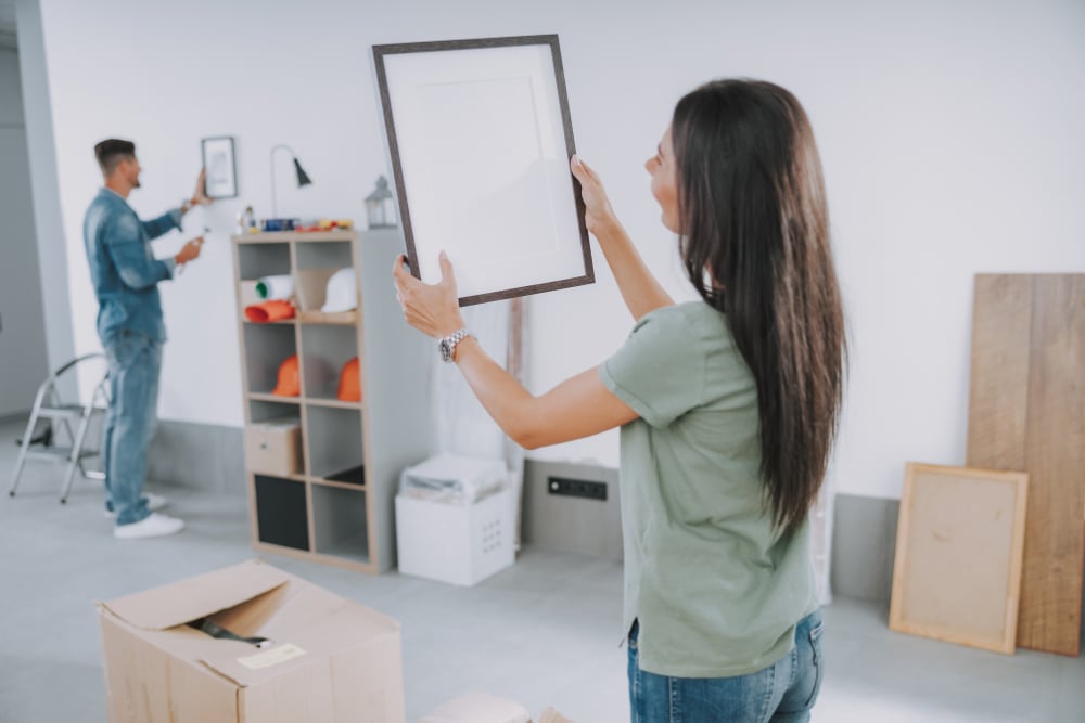 Two people pack a room, arranging framed pictures beside a cardboard moving box.