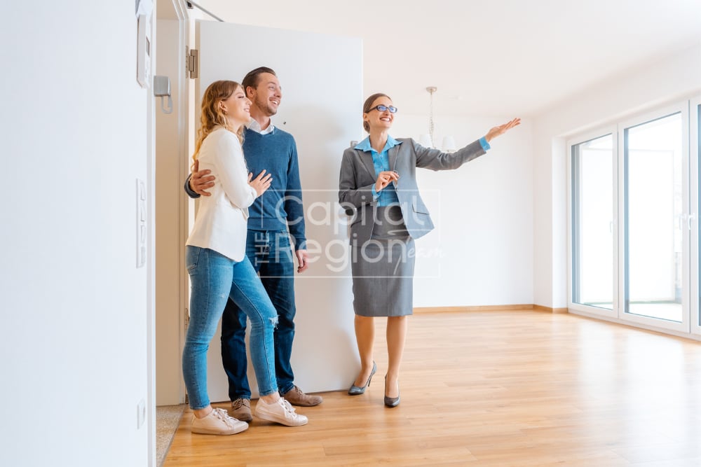 Agent shows a couple a bright vacant room with wood floors and large windows.