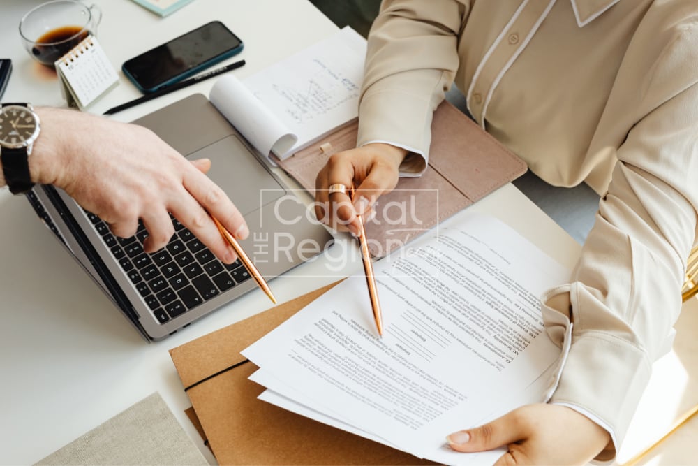 Two people review documents at a desk beside a laptop and notebook.