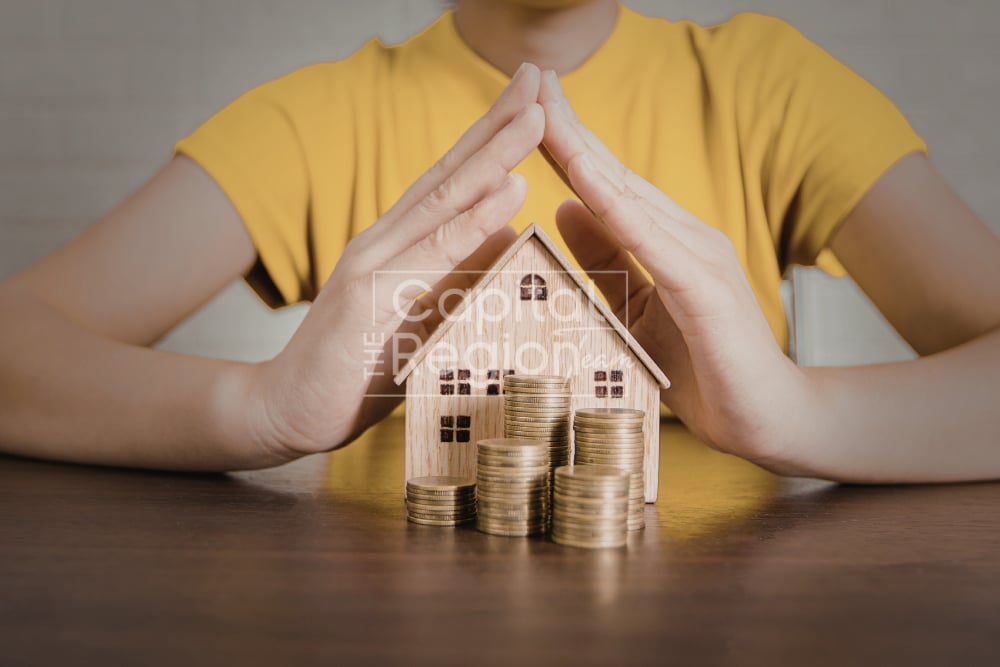 Hands shelter a model wooden house and stacks of coins.