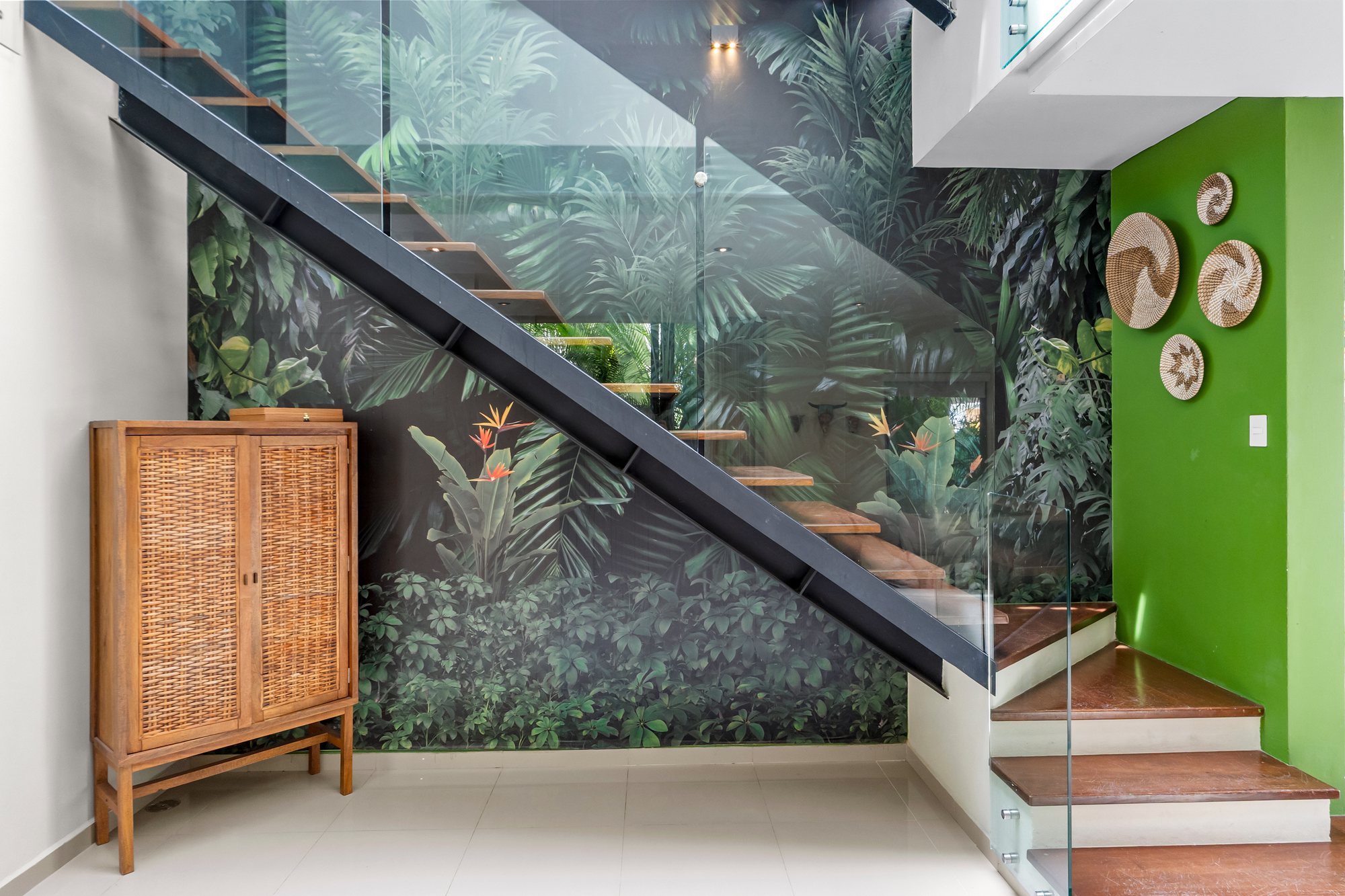 Escalera flotante de madera con barandal de cristal junto a un mural de selva