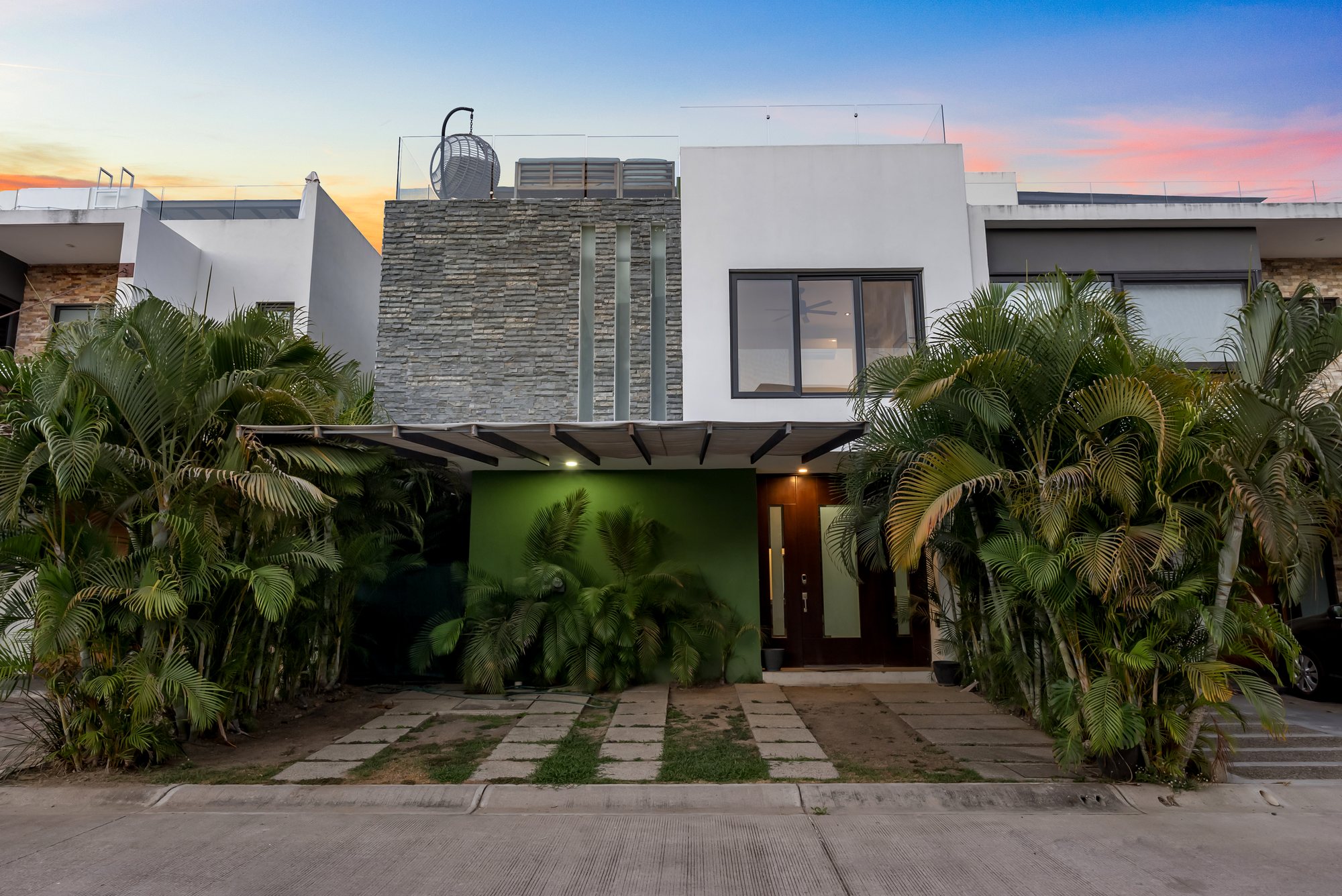 Fachada de la casa al anochecer, con palmeras y muro verde en la entrada