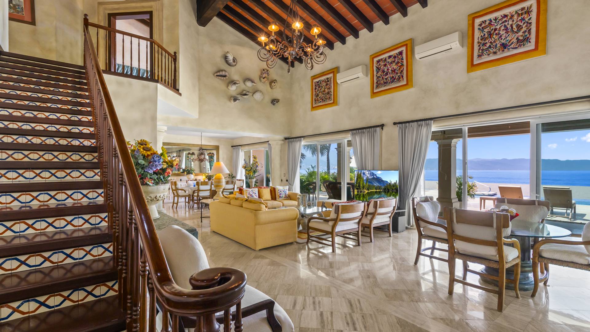 Double-height living room with wood-beam ceiling, open staircase and sliding doors to the ocean