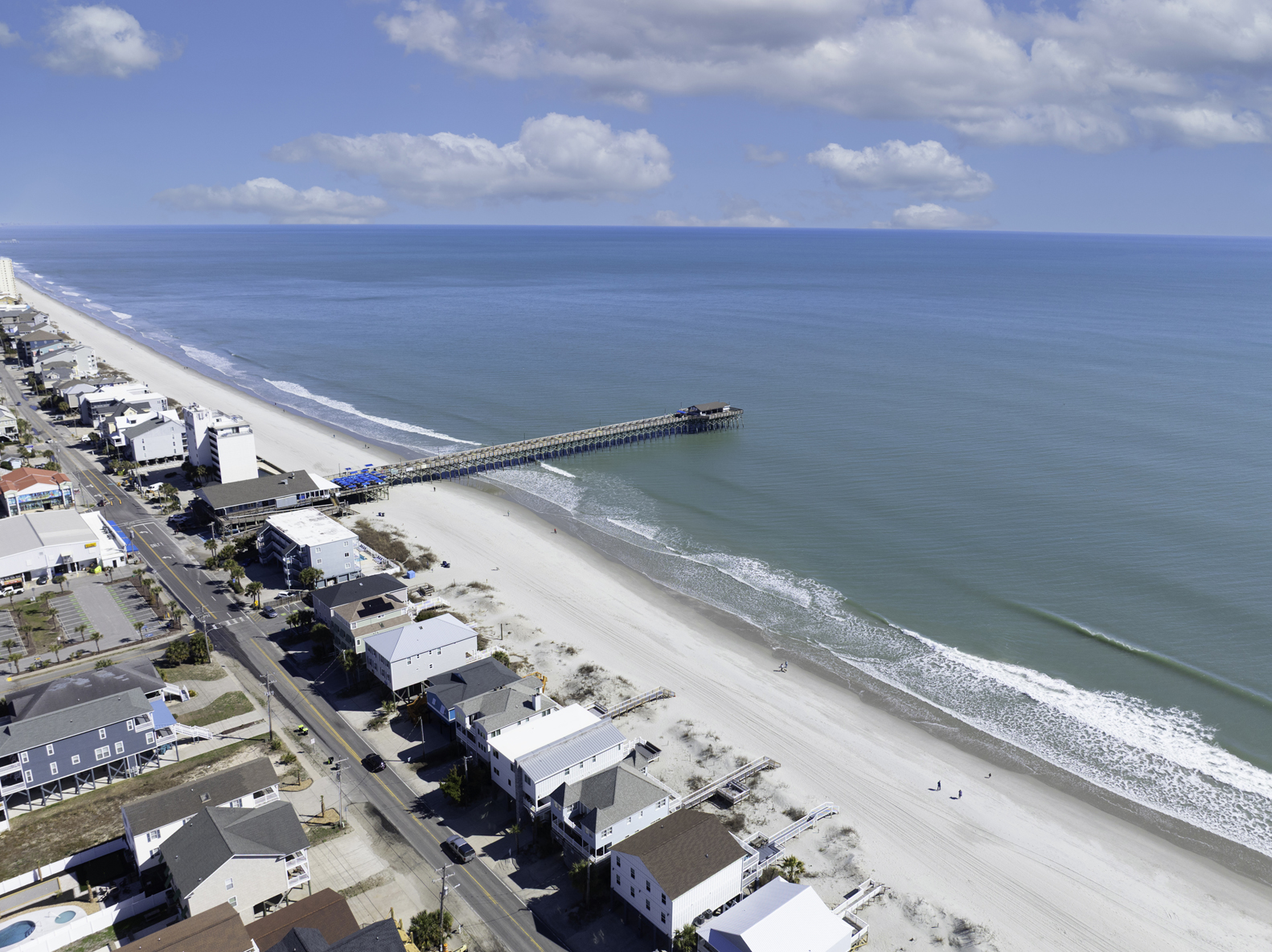Panoramic view of Garden City Beach, South Carolina featuring the Garden City Pier extending into the Atlantic Ocean with sandy shoreline, beachgoers, and oceanfront homes along the South Strand.