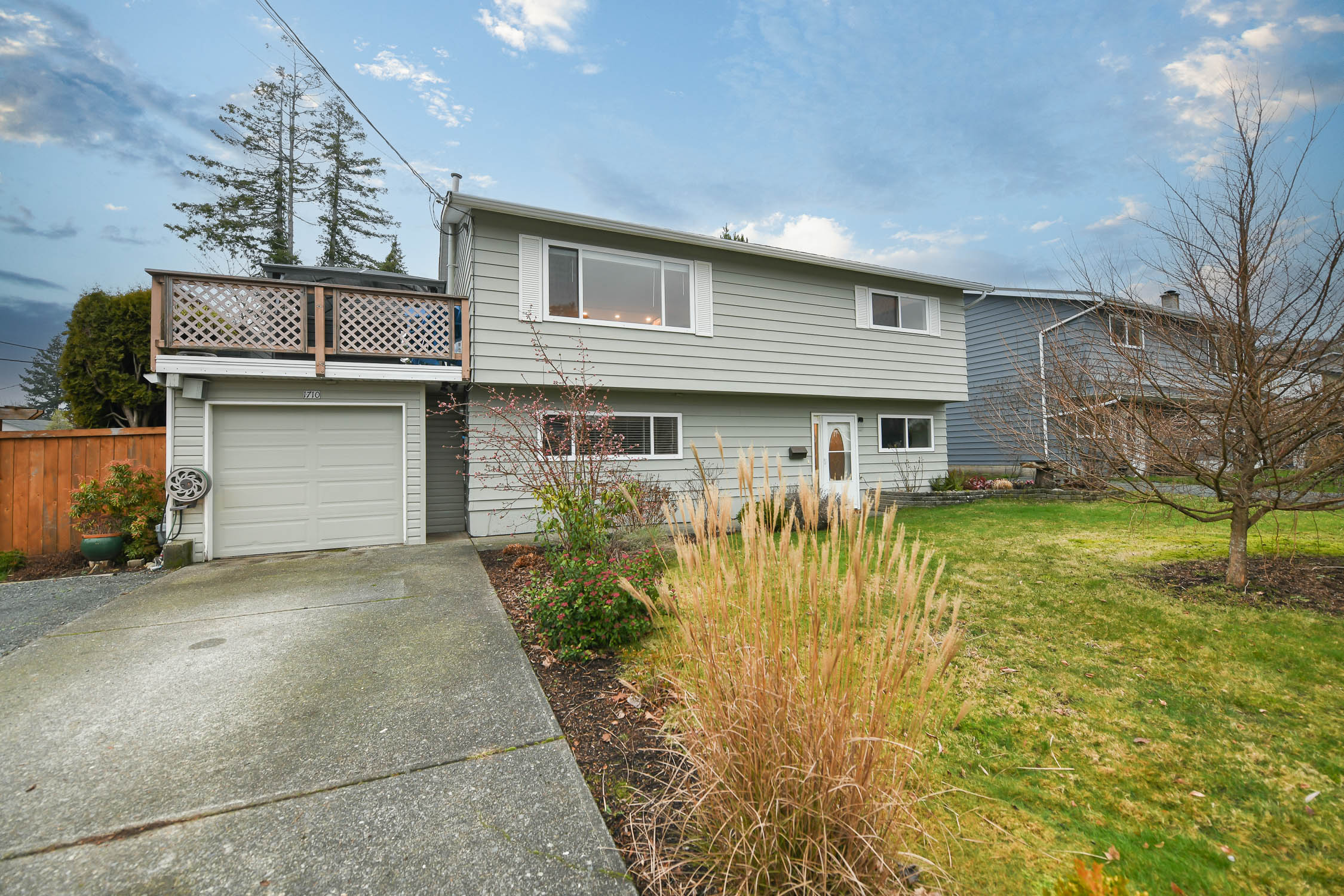 A spacious wrap-around sundeck overlooking a beautifully landscaped backyard with mature trees and greenery. In the distance, stunning mountain views stretch across the horizon beneath a bright, clear sky. The deck features sturdy railings and ample space for outdoor seating, capturing a peaceful, elevated view of both nature and the surrounding neighbourhood.
