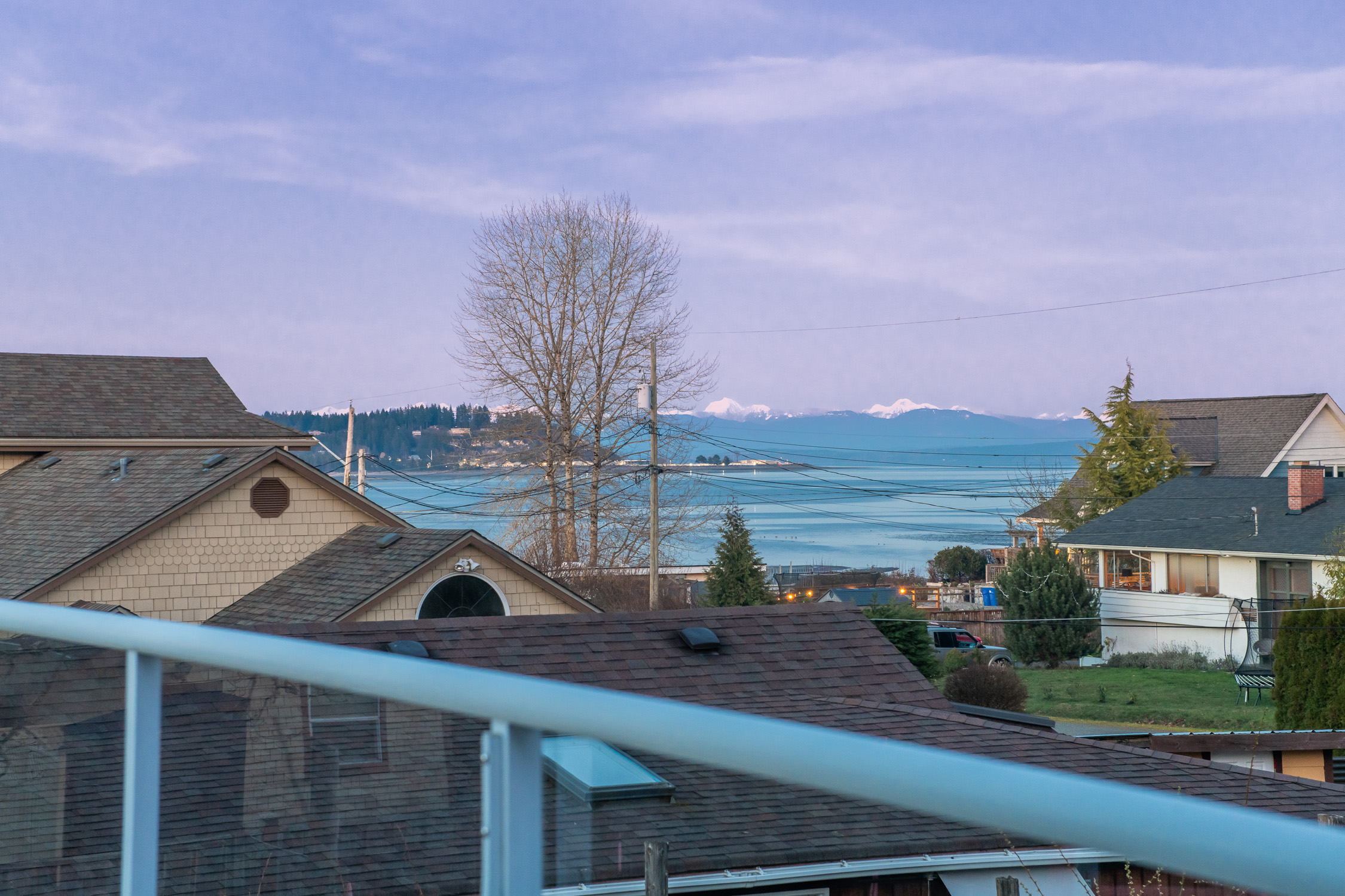 A spacious elevated deck overlooking the ocean, showcasing expansive coastal views across the Salish Sea. The vantage point captures stunning sunrises and the glow of Comox night lights in the distance, with the shoreline and water visible just steps away. The deck offers an ideal outdoor living space for relaxing or entertaining while enjoying the peaceful seaside setting and uninterrupted west coast scenery.