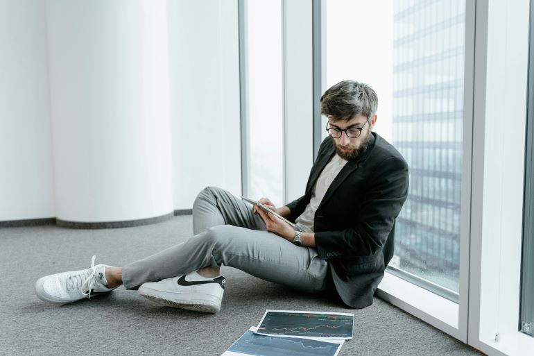 Man sitting on the floor near large windows, analyzing financial charts and graphs on a tablet