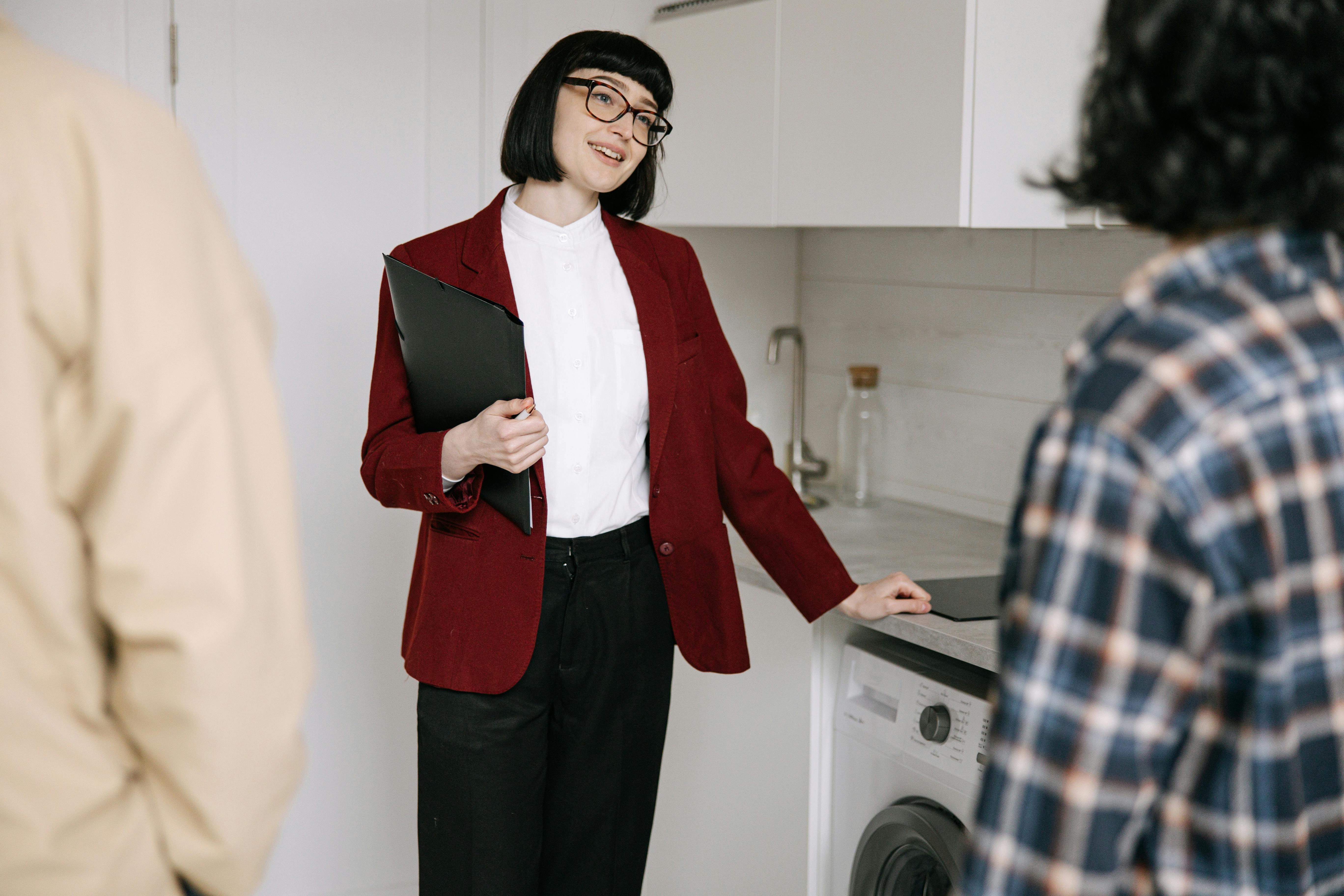 A real estate agent in a red blazer showing a laundry area to two clients inside a bright kitchen