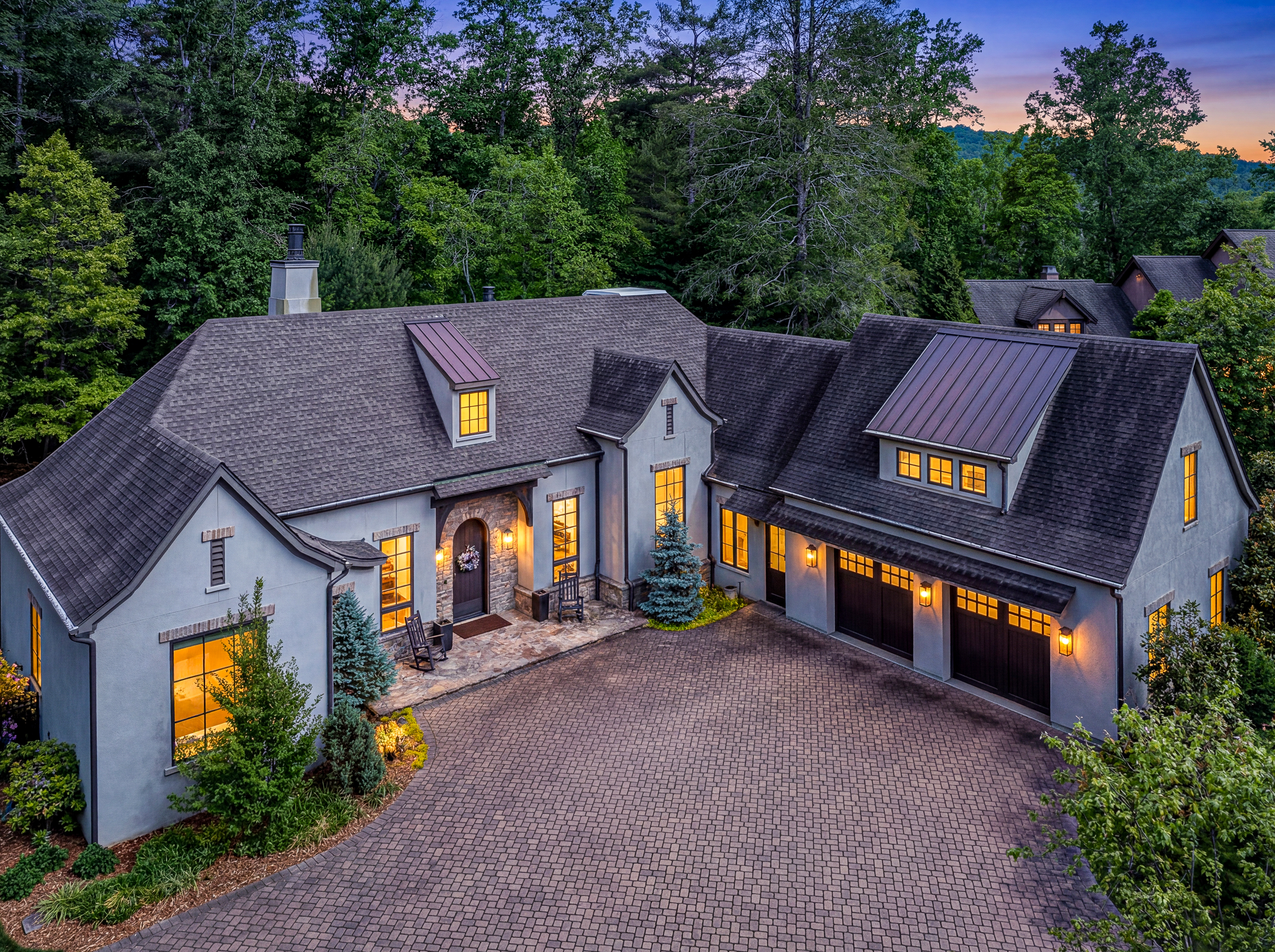 Luxury home at twilight in The Ramble Biltmore Forest, Asheville NC, featuring stone and stucco exterior, arched entry, paver driveway, and warm interior lighting surrounded by mature trees