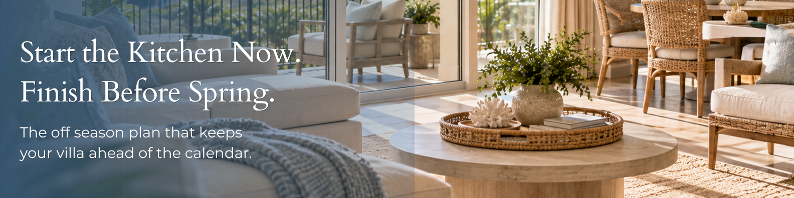 Sunlit villa living area with a cream sectional and knit throw in the foreground, a round stone-look coffee table styled with a woven tray, coral, and greenery, and rattan dining chairs beside glass doors opening to a balcony with outdoor seating